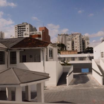 a foto mostra a rea do ptio do IPC, com rampas de acesso, coreto central, corredores e grandes janelas nos vrios prdios. O cho  cinza, as paredes so brancas e o telhado, marrom. Ao fundo, do lado de fora, h alguns prdios vizinhos. O cu est bem azul.