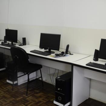 a foto mostra a sala de informtica. O piso  de taco marrom escuro. As mesas so brancas, lado a lado, prximas a uma parede branca. Os computadores so pretos, todos iguais, sobre as mesas. As cadeiras so pretas e ficam numa posio em que a pessoa senta de frente para a parede. Na foto, aparecem quatro computadores e duas cadeiras.