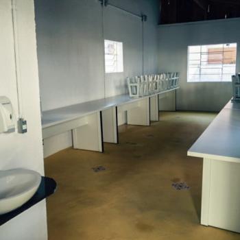 a foto mostra o refeitrio, com ngulo de quem o v da porta. Ao lado esquerdo est uma pequena pia branca e um aparelho para lcool gel. Ao fundo est o refeitrio, com duas mesas retangulares e longas, sendo uma de cada lado da parede (direita e esquerda). Os bancos brancos esto sobre as mesas, de cabea para baixo. As paredes so brancas e o piso  marrom.