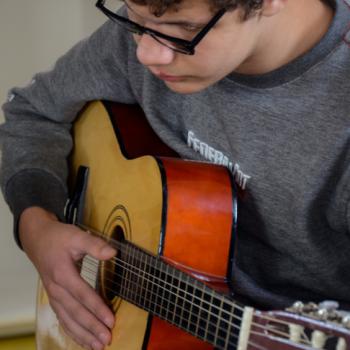 a imagem mostra um aluno sentado com um violo no colo. Tem sua mo direita nas cordas e a esquerda apoiada na base do instrumento musical. Ele olha para frente, tem a cabea baixa e est srio.
