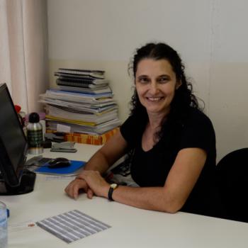 A foto mostra a diretora do CAEE Idamaris Singulani Costa e da Escola Osny Macedo Saldanha sentada  sua mesa,  direita da foto, olhando para foto e sorrindo. Ela est com os dois braos sobre a mesa, tendo a mo esquerda sobre a direita. Tem cabelos pretos e longos, semi-presos.