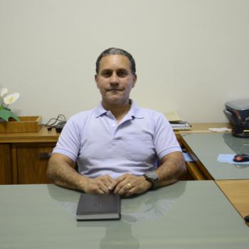 Foto de Carlos Roberto da Silva, coordenador do setor Financeiro e de Contabilidade. Ele est sentado  mesa, encostado a uma cadeira, com os dois braos sobre ela e as mos encostadas. Tem a sua frente, sobre a mesa,
uma agenda com capa preta. Ele tem cabelos pretos e um pouco grisalhos, est olhando para a foto com um leve sorriso. Usa uma camisa polo azul e relgio no pulso esquerdo.