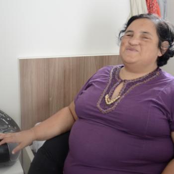 a imagem mostra uma moradora sentada  cama, ouvindo msica. Ela est com a cabea levemente erguida para cima, de olhos fechados e sorrindo enquanto coloca a mo direita sobre um rdio que est a sua frente, sobre um criado-mudo branco.