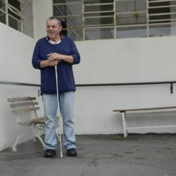 a imagem mostra um morador em p no ptio externo do IPC. Ele est sorrindo, com olhos fechados, a cabea ligeiramente virada para o lado direito. Ele est apoiado uma bengala branca, que segura em p com a mo direita. A mo esquerda est sobre a direita. Ele est prximo a uma parede branca, com bancos e janelas atrs dele.