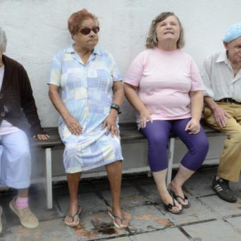 a imagem mostra trs moradoras e um morador sentados no banco na rea externa do IPC, lado a lado, num momento de convvio e descontrao. A senhora da direita est olhando para baixo e sorrindo, tendo as duas mos sobre o banco, ao lado do corpo.  esquerda dela (direita da foto), uma senhora usa culos de sol e olha para a frente. Est sria, com as mos sobre as pernas.  esquerda dela (direita da foto), outra senhora sorri enquanto olha para cima.  esquerda dela (direita da foto), um senhor tambm sorri. Ele usa um gorro azul na cabea.