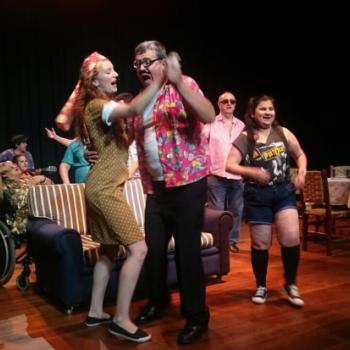 a imagem mostra uma cena da pea teatral, com dois atores danando em par e cantando, ao mesmo tempo. Atrs deles h um sof listrado. Atrs e ao lado do sof, h outros atores, que tambm esto cantando. Uma atriz est sentada em cadeira de rodas. Ao fundo, outro toca violo. Do lado direito da foto, ao fundo, h uma retangular mesa para refeies, com trs cadeiras, xcaras e jarra de bebida em forma de abacaxi.