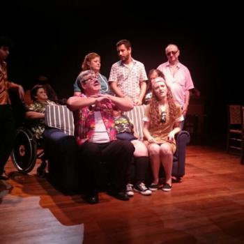 a imagem mostra uma cena da pea teatral, com dois atores sentados num sof listrado, gesticulando e falando. Atrs do sof, h outros atores em p, em silncio, srios. Uma atriz est sentada em cadeira de rodas.