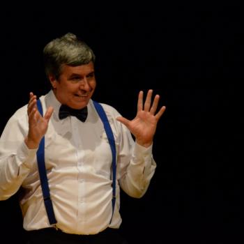 a imagem mostra uma cena da pea teatral, com recorte em um ator sorrindo enquanto fala, com as duas mos erguidas (braos dobrados na altura do cotovelo, na cintura).
