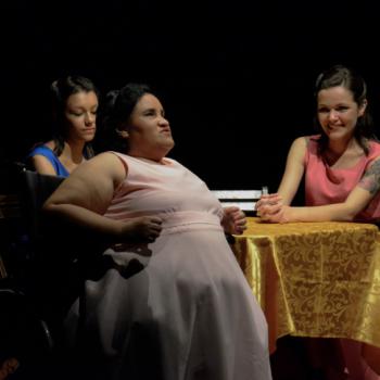 a imagem mostra uma cena da pea teatral, com trs atrizes sentadas a uma mesa com toalha dourada. A atriz principal na cena est falando, sentada numa cadeira de rodas. Do lado esquerdo dela, a atriz est olhando para baixo. E do lado direito dela, uma outra atriz sorri e olha para a atriz que est falando.